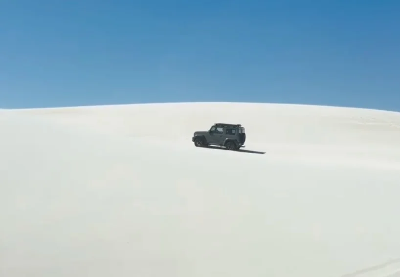 Suzuki Jimny crossing white sand dunes beneath a blue sky
